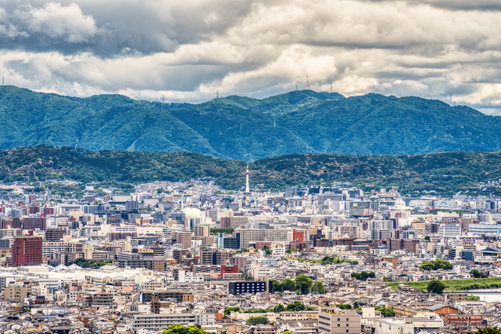 Die Aufnahme zeigt die dichte Stadtlandschaft von Kyoto mit vielen Gebäuden und bewaldeten Bergen unter wolkigem Himmel.