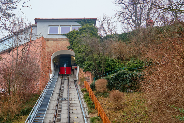 Die rote Schloßbergbahn fährt auf Schienen steil den grünen Hang hinauf in Richtung einer Tunnelöffnung.