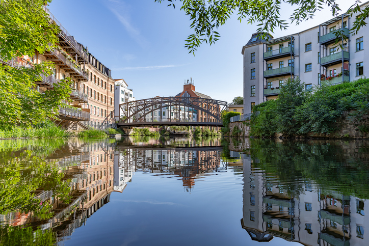 Ein ruhiger Kanal in Leipzig, gesäumt von Backsteingebäuden und einer Eisenbrücke, die sich im Wasser spiegeln.