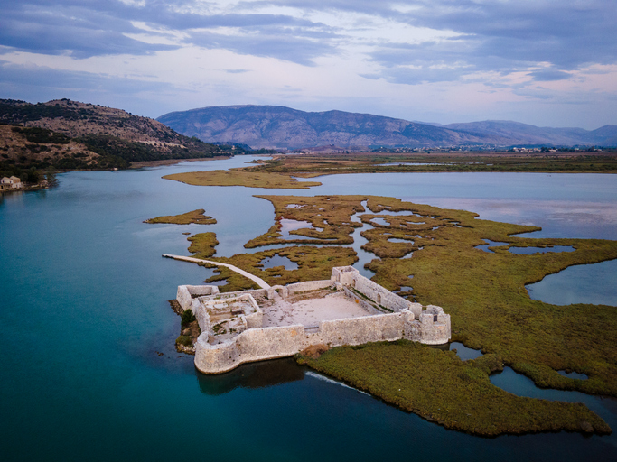 Eine historische Steinanlage liegt zwischen Wasserarmen und flachen grünen Uferflächen in weiter Landschaft.
