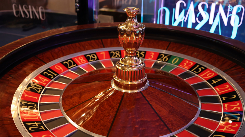 Eine Nahaufnahme eines glänzenden hölzernen Roulette-Rades in einem Casino, im Hintergrund leuchtet ein Neonschild mit der Aufschrift „Casino“.