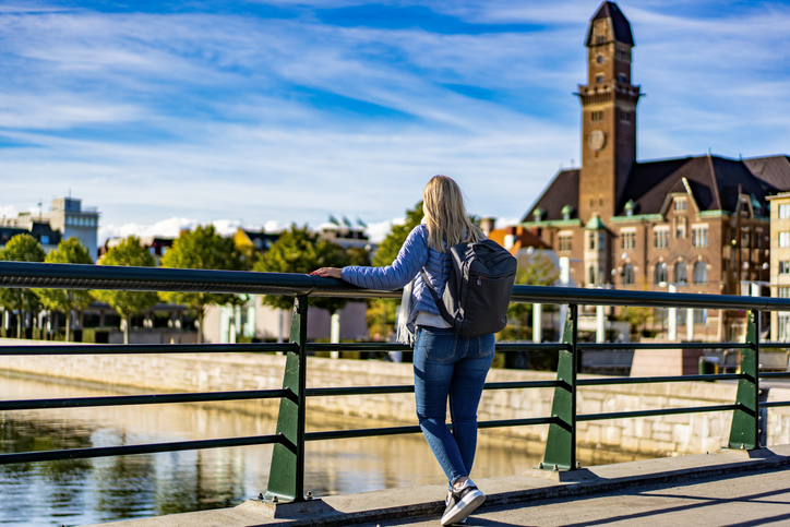 Eine Frau mit Rucksack lehnt an einem Geländer und blickt über das Wasser auf das markante Rathaus von Malmö.