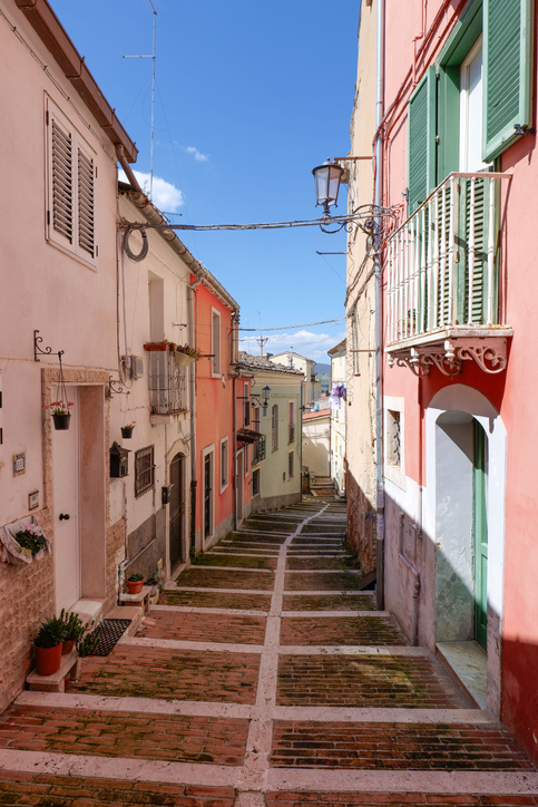 Sonnige, gepflasterte Steintreppe zwischen pastellfarbenen Häusern mit grünen Fensterläden und Balkonen in einer historischen Gasse.