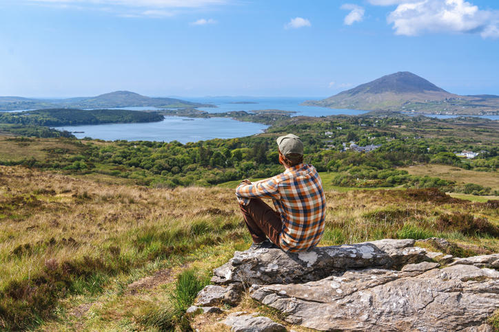 Ein Wanderer sitzt auf einem Felsen und blickt über die weite Hügellandschaft und Meeresbuchten der Westküste.