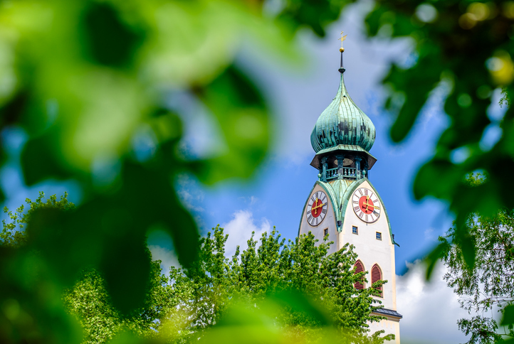 Nahaufnahme des hellen Kirchturms mit seiner grünen Zwiebelkuppel, eingerahmt von grünen Blättern.