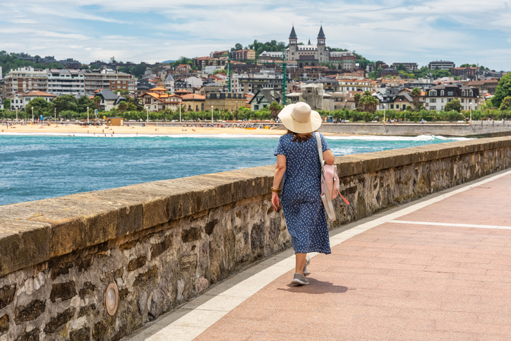 Eine Frau geht an der Uferpromenade entlang mit Strand und Stadt im Hintergrund.