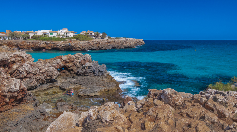 Eine felsige Küstenbucht mit türkisblauem Wasser und wenigen Häusern auf den Klippen liegt unter tiefblauem Himmel.