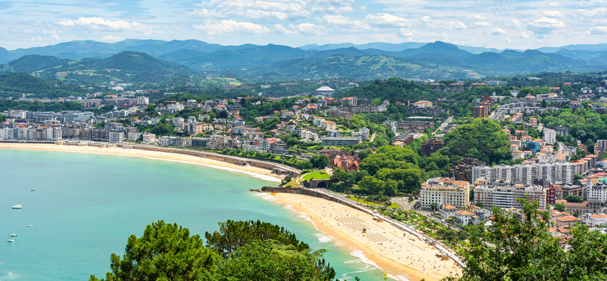 Eine weite Küstenansicht zeigt Strand, Stadt und grüne Hügel aus der Höhe.