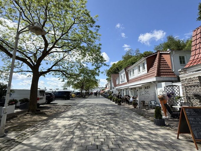 Eine gemütliche gepflasterte Straße mit Cafés und Fachwerkhäusern, die von grünen Bäumen gesäumt wird.