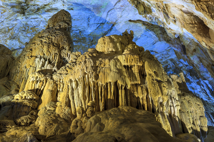Riesige, beleuchtete Stalaktiten und Stalagmiten in einer monumentalen Höhle unter einer bläulich schimmernden Decke.