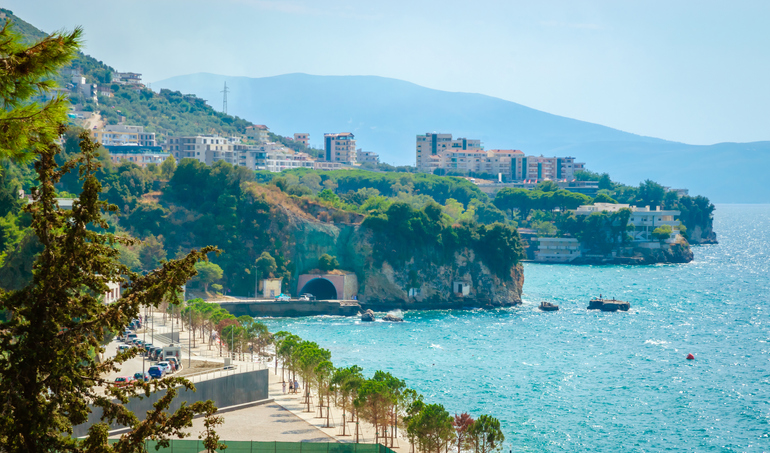 Eine Küstenstraße, Felsen und Gebäude ziehen sich entlang des glitzernden Meeres vor bergiger Kulisse.