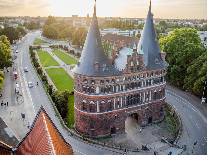 Eine beeindruckende Ansicht des massiven Holstentors aus Backstein, das leicht geneigt auf einem grünen Platz steht, während die Sonne untergeht.