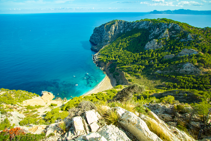 Eine abgelegene Sandbucht liegt unter steilen grünen Felsen am tiefblauen Meer.