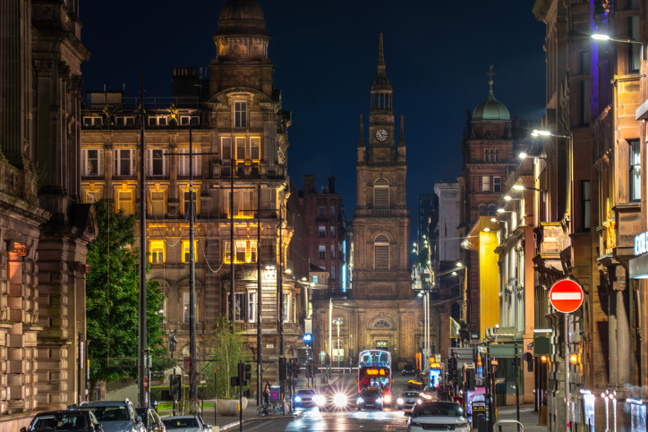 Eine nächtliche Straßenansicht mit historischen Sandsteingebäuden, die auf den beleuchteten Kirchturm der St. George’s Tron Church zuführt.