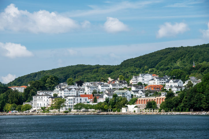 Ein Blick vom Wasser auf weiße Villen im Bäderstil, die an einem bewaldeten Hang direkt an der Küste liegen.