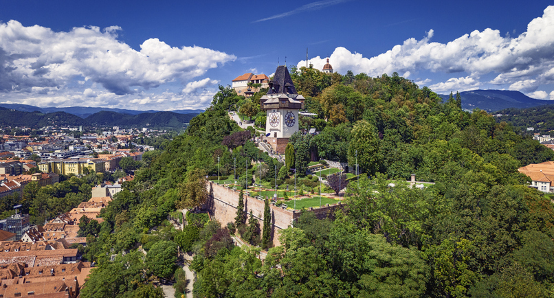 Eine Luftaufnahme des dicht bewaldeten Schloßbergs mit dem markanten Uhrturm unter strahlend blauem Himmel.