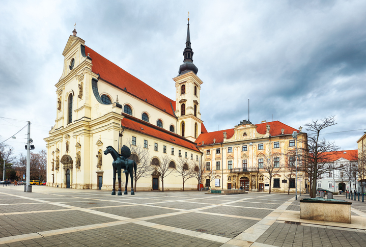 Eine helle Kirche mit Turm steht an einem weiten fast leeren Platz.