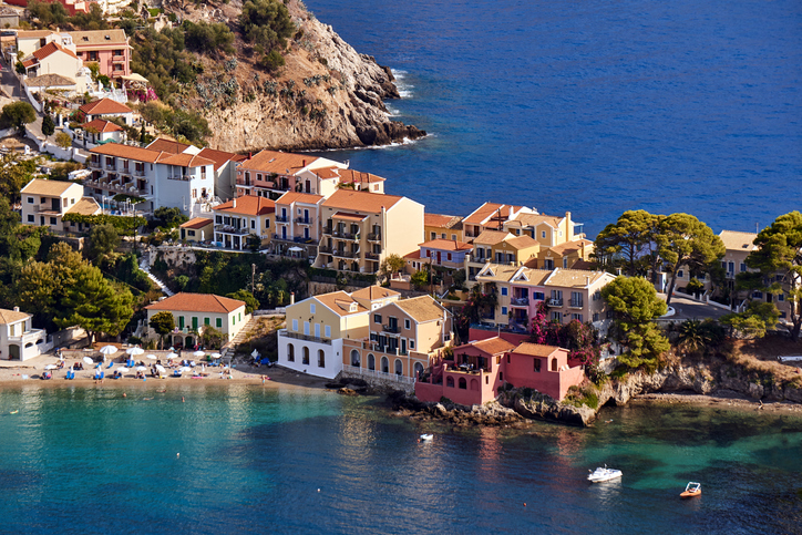 Bunte Häuser schmiegen sich an eine schmale Landzunge zwischen zwei tiefblauen Meeresbuchten unter strahlender Sonne.