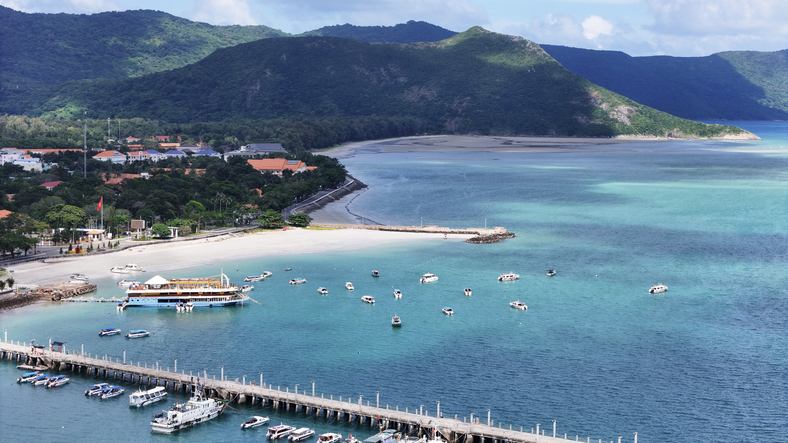 Luftaufnahme einer türkisfarbenen Bucht mit weißem Sandstrand, zahlreichen Booten im Wasser und einem langen Pier im Vordergrund.