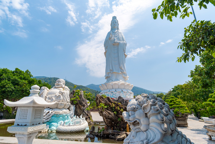 Eine Nahaufnahme einer weißen Wächterlöwen-Statue im Vordergrund, dahinter eine sitzende Buddha-Figur und die hoch aufragende Lady Buddha Statue.