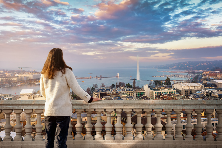 Eine Frau blickt von einer Terrasse auf Stadt, See und Wasserfontäne.