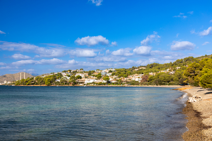 Eine stille Küstenlinie mit flachem Wasser, Kiesstrand und Häusern an einem grünen Hang liegt unter blauem Himmel.