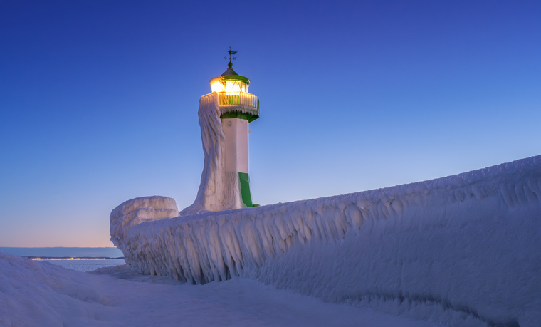 Ein grün-weißer Leuchtturm und die Hafenmauer sind nach einem Wintersturm vollständig mit bizarren Eiszapfen überzogen.