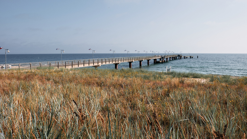 Eine lange Holzbrücke führt weit hinaus auf die offene Ostsee, umgeben von Dünengras und sanften Wellen.