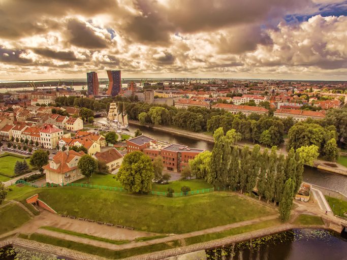 Eine weite Stadtansicht zeigt Grünflächen, Wasserwege und Dächer unter dichten Wolken.