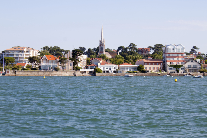 Über dem Wasser sind Villen, eine Uferlinie und ein schmaler Kirchturm in der Ferne zu sehen.