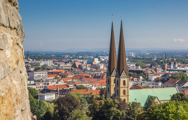 Zwei hohe Kirchtürme ragen vor einem Stadtpanorama mit roten Dächern und Bäumen auf.