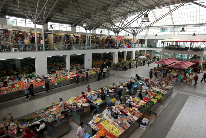Obst, Gemüse und Stände füllen eine helle Markthalle mit vielen Menschen.