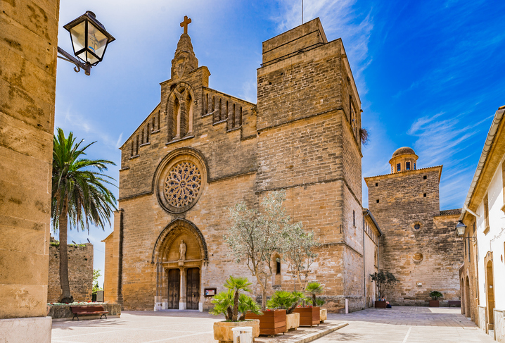 Die massive, festungsartige Fassade der Kirche Sant Jaume in Alcudia mit einer großen Rosette unter strahlend blauem Himmel.