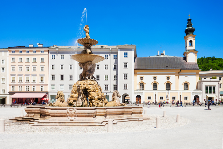 Ein großer Brunnen steht vor hellen Häusern und einer Kirche unter blauem Himmel.