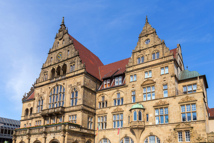 Ein großes historisches Gebäude mit reich verzierter Fassade, Uhr und roten Dächern steht unter blauem Himmel.