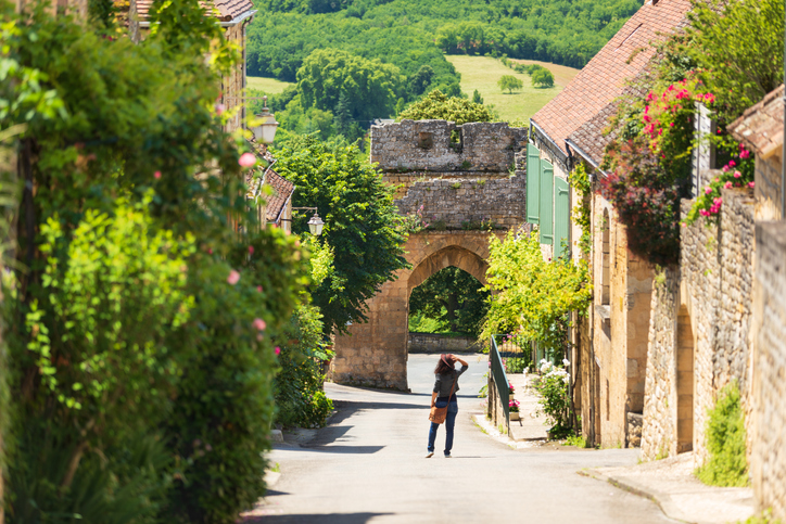 Eine ruhige Straße mit Natursteinhäusern, Blumen und einem alten Torbogen führt durch ein grünes Dorf.