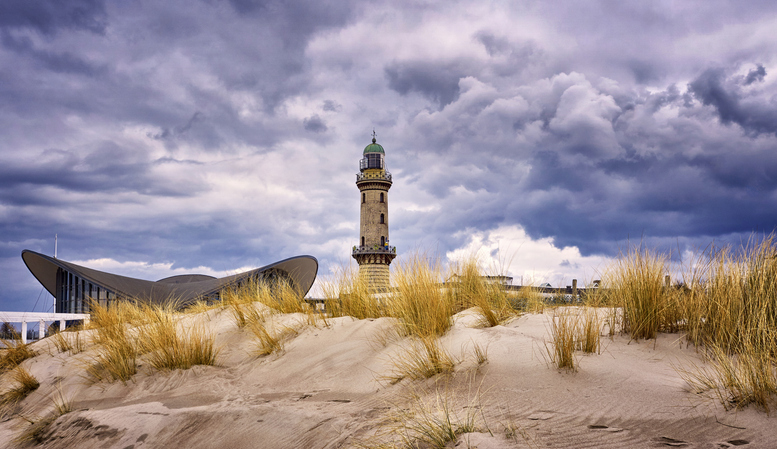 Der markante Leuchtturm von Warnemünde und das geschwungene Dach des Teepotts hinter sandigen Dünen unter einem dramatischen Wolkenhimmel.