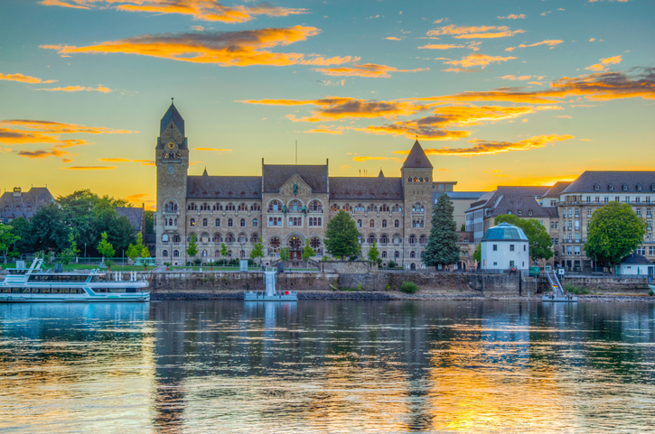 Ein prächtiges, schlossähnliches Gebäude am Rheinufer mit einem markanten Uhrturm im warmen Licht des Abendhimmels.