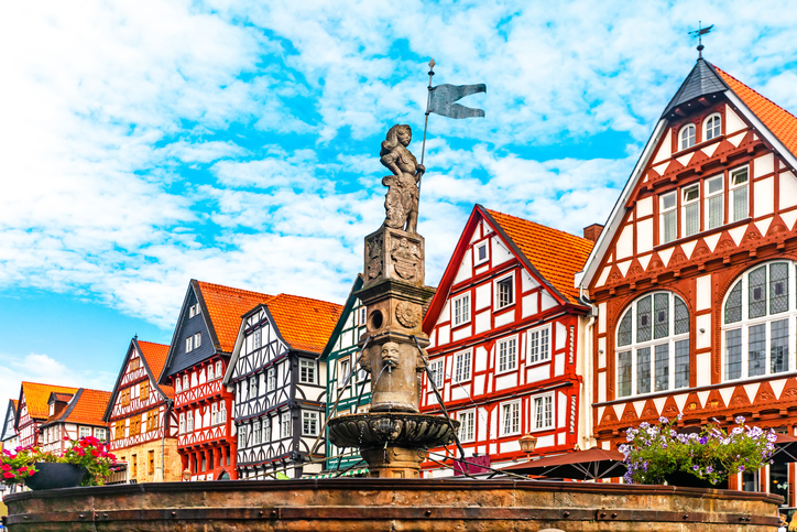Ein kunstvoller Steinbrunnen mit einer Ritterstatue vor einer Reihe prachtvoller rot-weißer Fachwerkhäuser unter blauem Himmel.