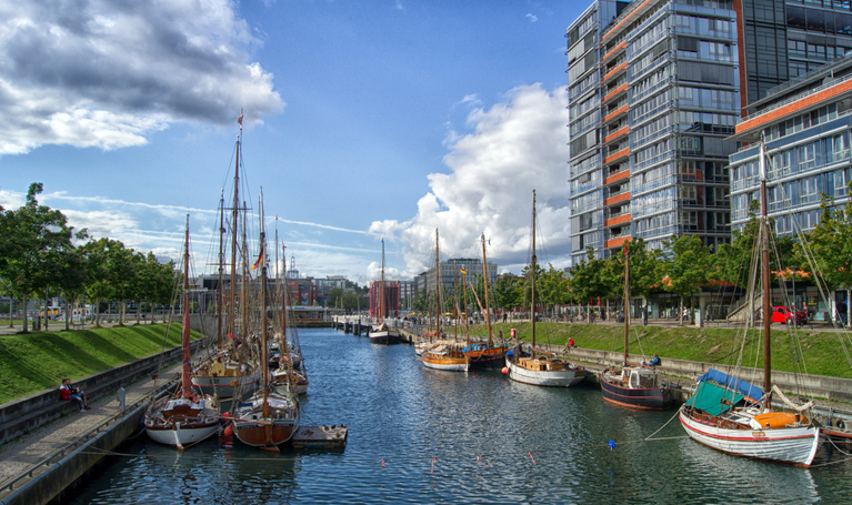 Traditionelle Segelboote liegen im Hafen von Kiel vor modernen Gebäuden und blauem Himmel.