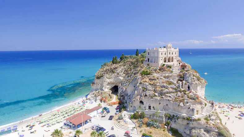 Luftaufnahme der auf einem Felsen thronenden Kirche Santa Maria dell'Isola in Tropea, umgeben von einem feinen Sandstrand und kristallklarem, blauem Meer.