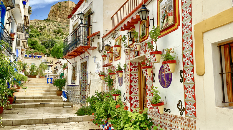 Eine malerische, mit Blumen geschmückte Gasse im traditionellen Viertel Santa Cruz in Alicante mit Blick auf die Burg