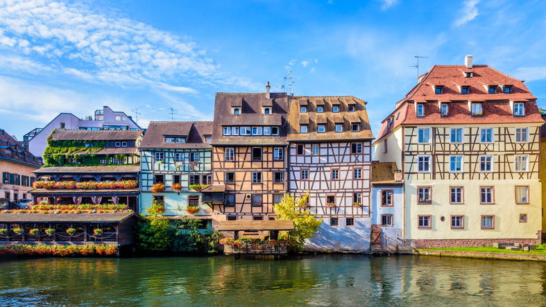 Eine Reihe historischer Fachwerkhäuser in verschiedenen Farben direkt am Wasser der Ill unter blauem Himmel.
