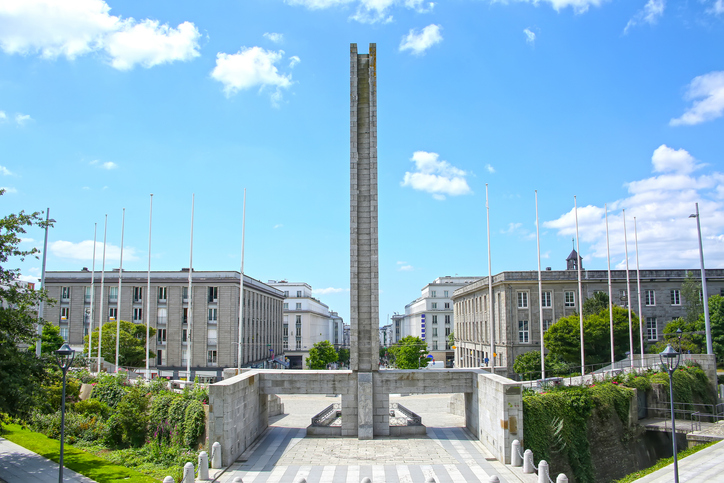 Ein hoher, schmaler Steinturm steht im Zentrum eines großen Platzes, flankiert von symmetrischen Gebäuden und Fahnenmasten.