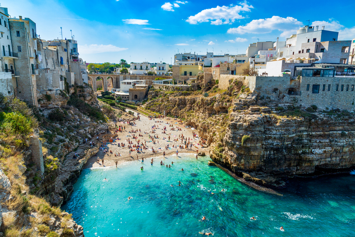 Ein sonniger Tag in der berühmten Bucht von Polignano a Mare mit vielen Badegästen am Strand und im glasklaren, azurblauen Wasser der Adria.