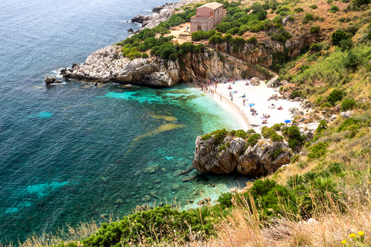 Eine kleine, versteckte Bucht mit weißem Sandstrand und türkisblauem Wasser bei San Vito Lo Capo, umgeben von felsigen Klippen und grüner Vegetation.