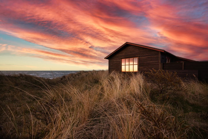 Eine kleine Holzhütte steht inmitten von hohen Dünengräsern bei einem dramatischen Sonnenuntergang in Hirtshals