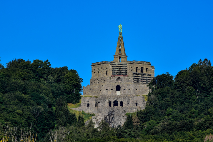 Das massive Steingebäude des Herkules-Monuments auf einem bewaldeten Hügel, gekrönt von der grünen Herkules-Statue.