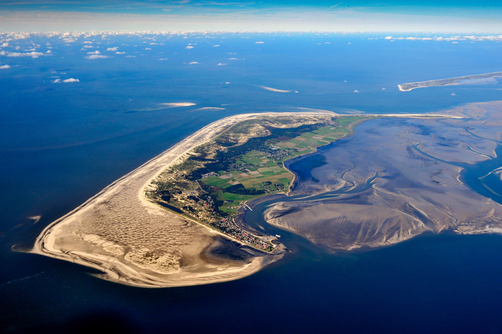 Luftbild der Insel Amrum mit ihrem breiten, hellen Sandstrand, den grünen Dünenlandschaften und dem angrenzenden Wattenmeer unter blauem Himmel.