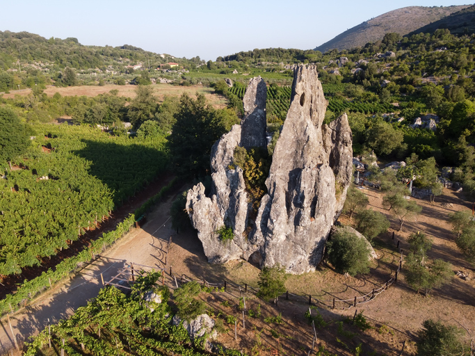 Eine gewaltige, zerklüftete Felsnadel ragt inmitten von Weinbergen und Olivenhainen in einer hügeligen Landschaft empor.